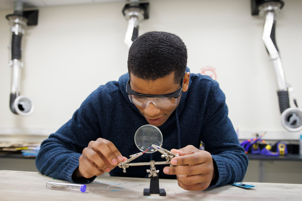 An Indiana University student in a lab uses a magnifying glass to work with small tools.