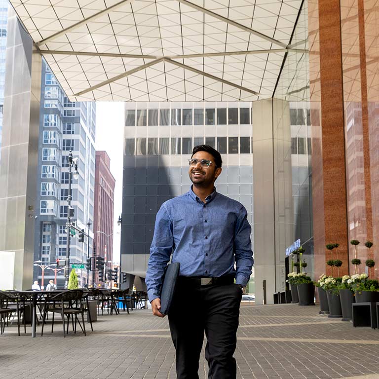 A male student walks through a glass corridor while admiring the space around him, tall city buildings stand in the background.