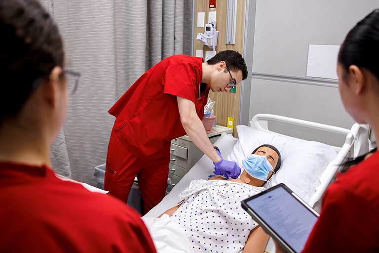 Two nursing students watch a male nursing student attend to a medical manikin in a hospital simulation. 