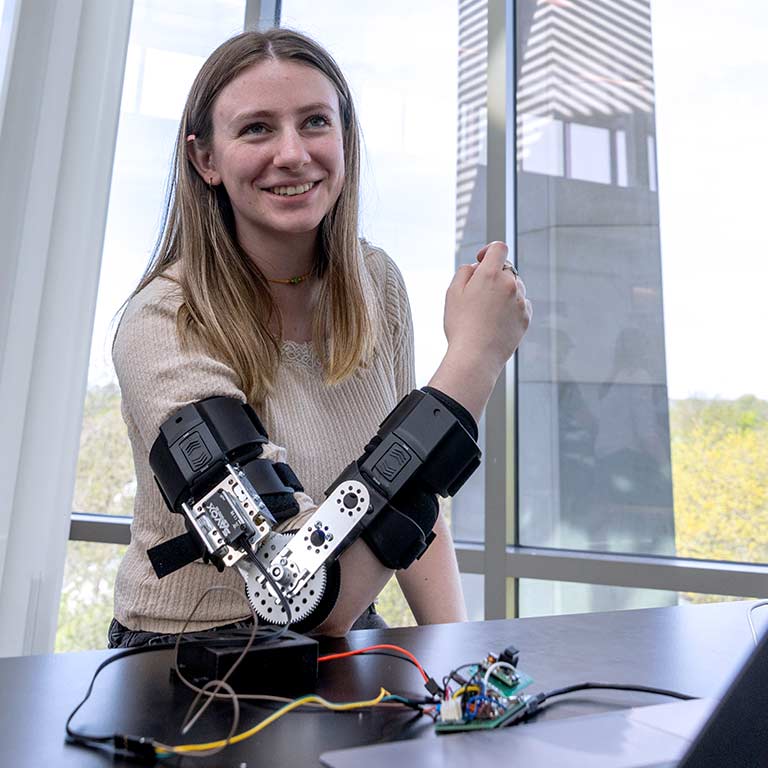 A female student smiles as she shows a robotic arm.