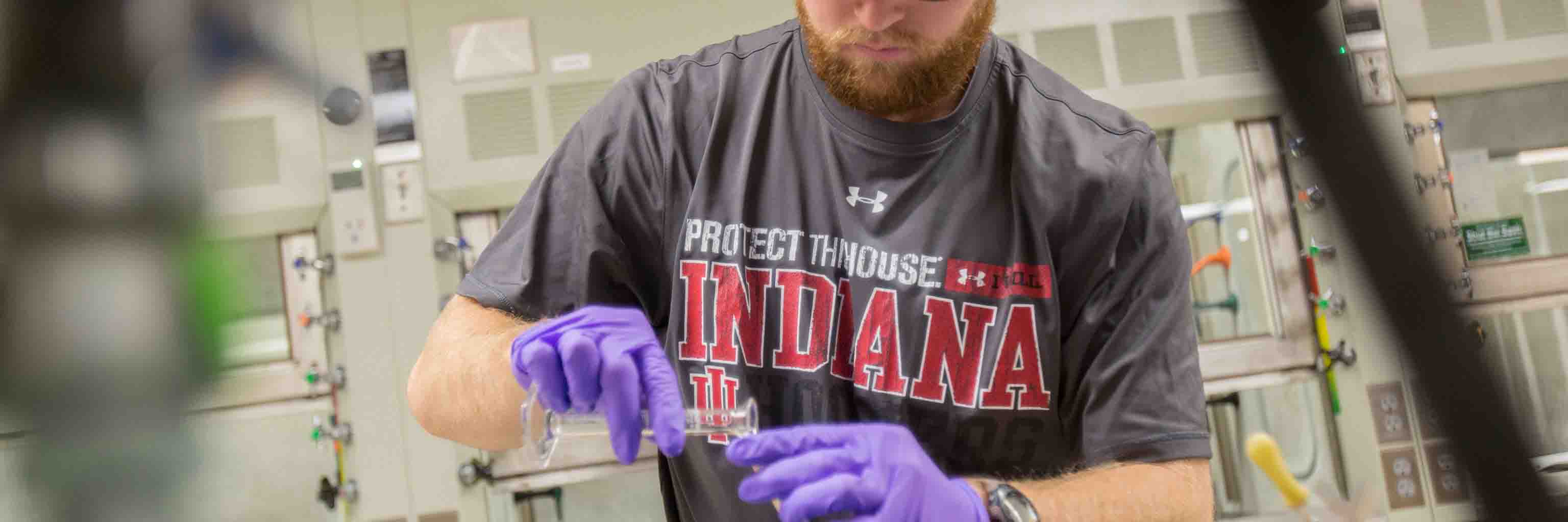 A student wearing purple gloves pours something from one test tube to another.