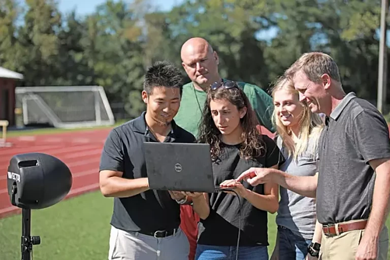 Concussion researcher Kei Kawata and colleagues are on an outdoor athletic field looking at a laptop.