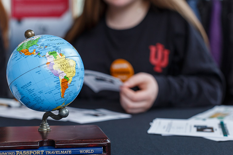 A small globe sits on a table. A student wearing an IU shirt is in the background.