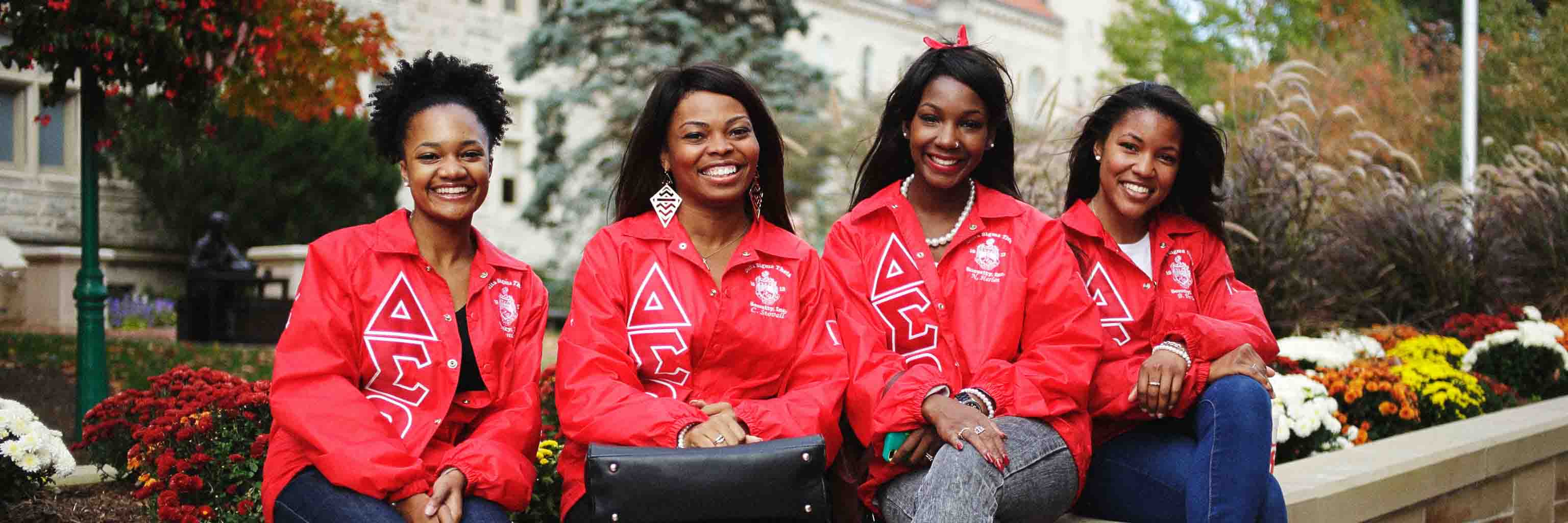 Four African American women in sorority jackets sit smiling on a bench.